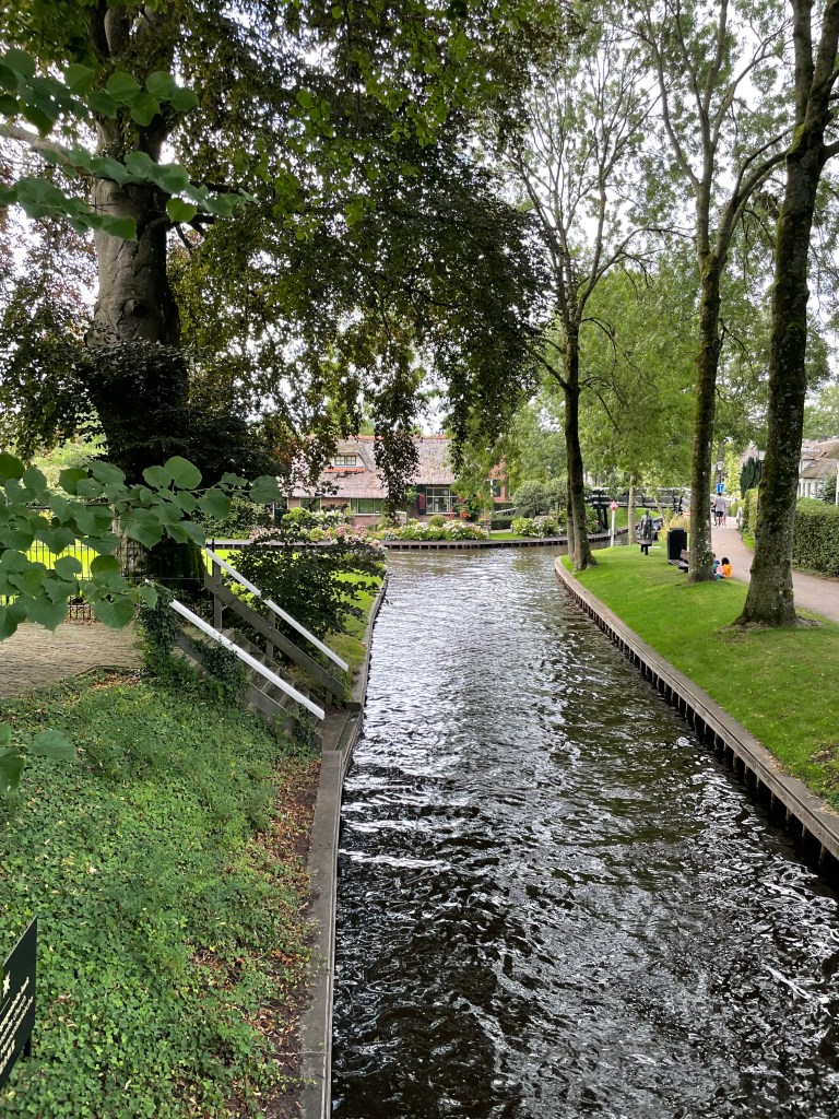 Photo of canal with homes bordered by gardens and walking paths with people in the background.