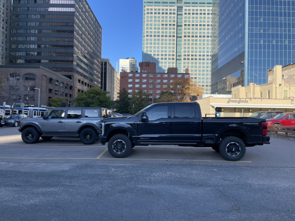 Cars are parked in a parking lot with large buildings in the background. There is a dark pick-up truck that is so large it doesn't fit in its parking spot.