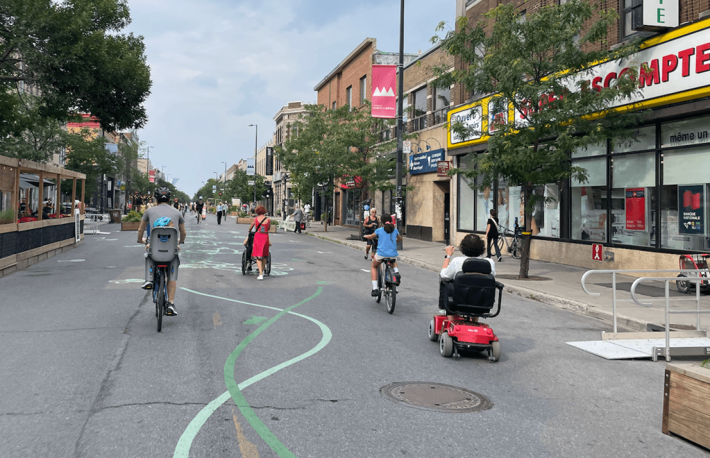 A street closed off to cars. People of a wide range of ages are biking, riding wheelchairs, and walking. There are shops bordering the sidewalk on one side of the street and there is restaurant seating blocked off on the other side.