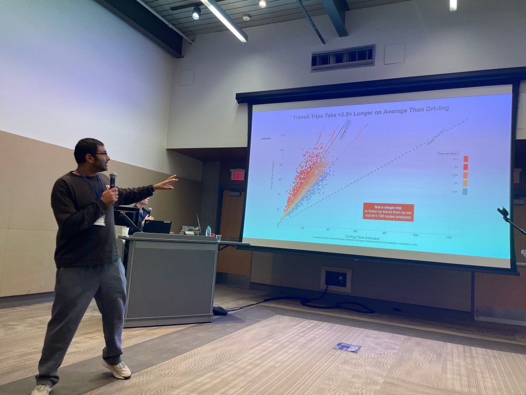 A man in a conference room points to a PowerPoint slide with a scatterplot depicting differences in travel times, with driving time on the X axis and transit time on the Y axis.