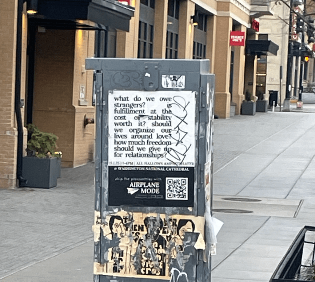 A metal utility box is in the middle of a sidewalk with storefronts behind it. On the utility box are large stickers, including one that reads: "what do we owe strangers? is fulfillment at the cost of stability worth it? should we organize our lives around love? how much freedom should we give up for relationships?"