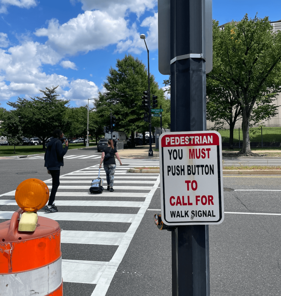 In the background, two people walk across a street in a zebra crosswalk. In the foreground, a sign on a post reads: "Pedestrian you must push button to call for walk signal".