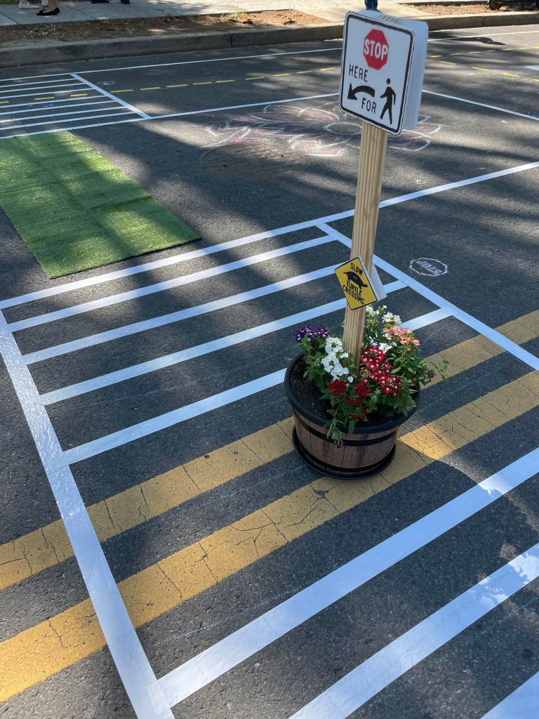 A miniature "stop here for pedestrians" sign planted in a pot of flowers and placed in the middle of a crosswalk.