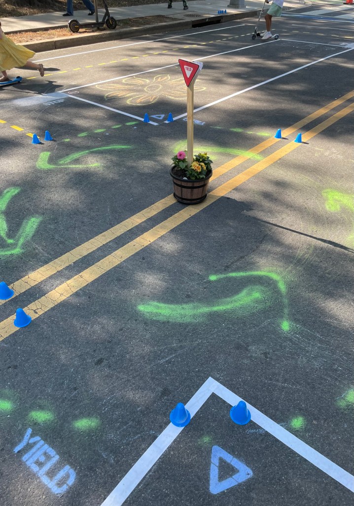 A miniature traffic circle with a yield sign in the middle, with two children riding scooters in the background and chalk art on the pavement.