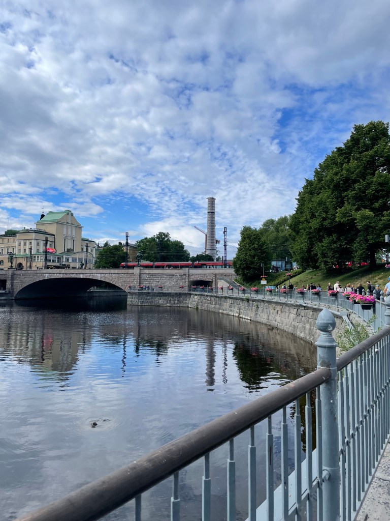 A riverbank is lined with a sidewalk, with flowers planted in pots on the railing and old buildings are behind an arched bridge in the background.