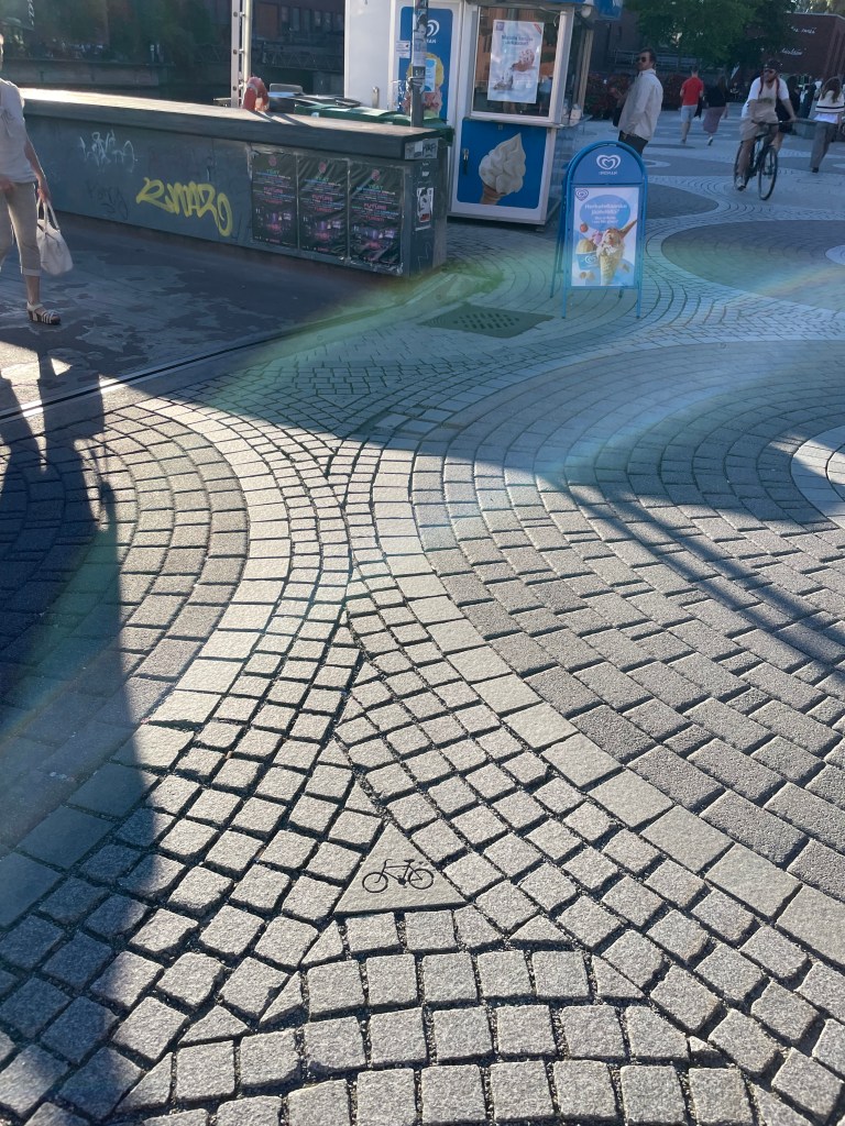 A sidewalk has gray pavers - one of the pavers is a triangle with a bicycle. 