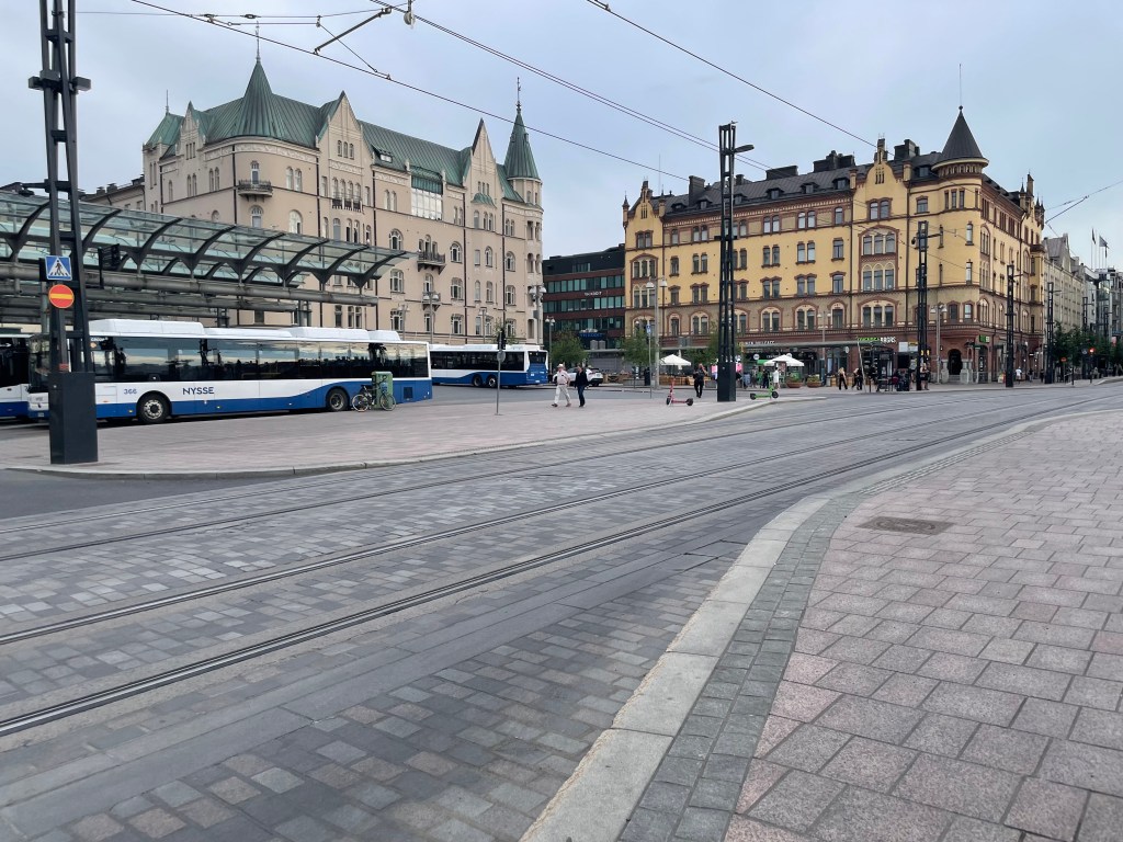 An old city center has an outdoor bus terminal in the background. Tram lines are in the street in the foreground. 