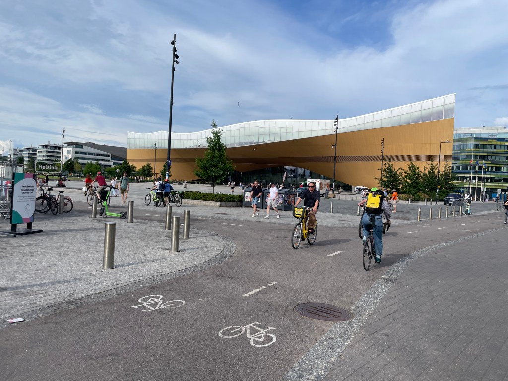 A large yellow library is in the background, a bike path is in the foreground as people riding bikes pedal along it. 