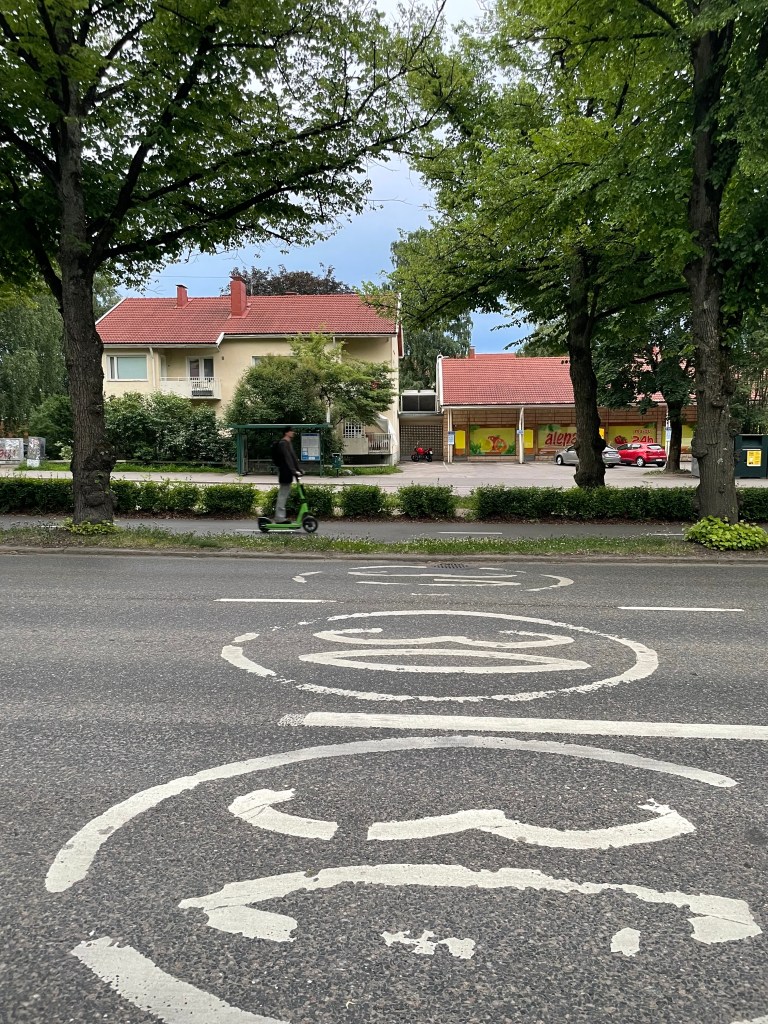A 30 km marking is painted on a three lane street, with someone riding a scooter on a center-running multi-use path in the background. 
