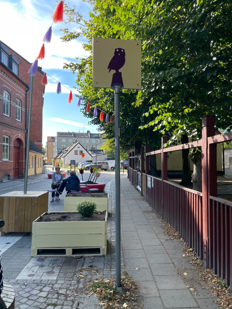 A street is closed to cars with the addition of planters, benches, and street art. An owl is painted on a street sign. Two kids sit and talk. 