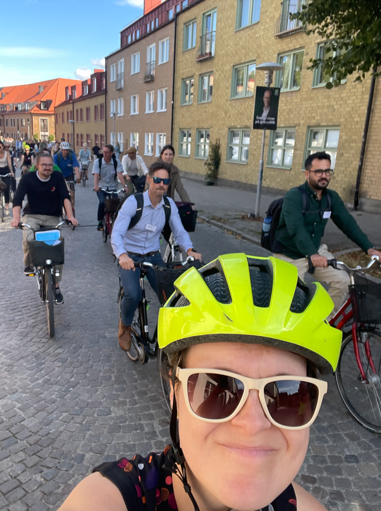 A woman wearing a yellow helmet taking a selfie of herself and a group of people behind her, all riding bicycles on a cobble-stone street.