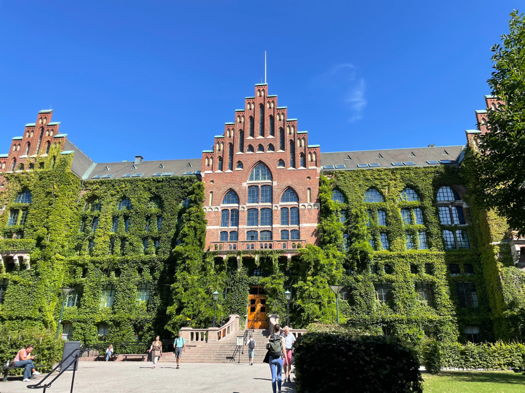The photo is taken from the outside of a large brick building (a library) covered with ivy and greenery. Behind the building is a blue sky in the background; in the foreground people are walking up to the building or sitting on benches.